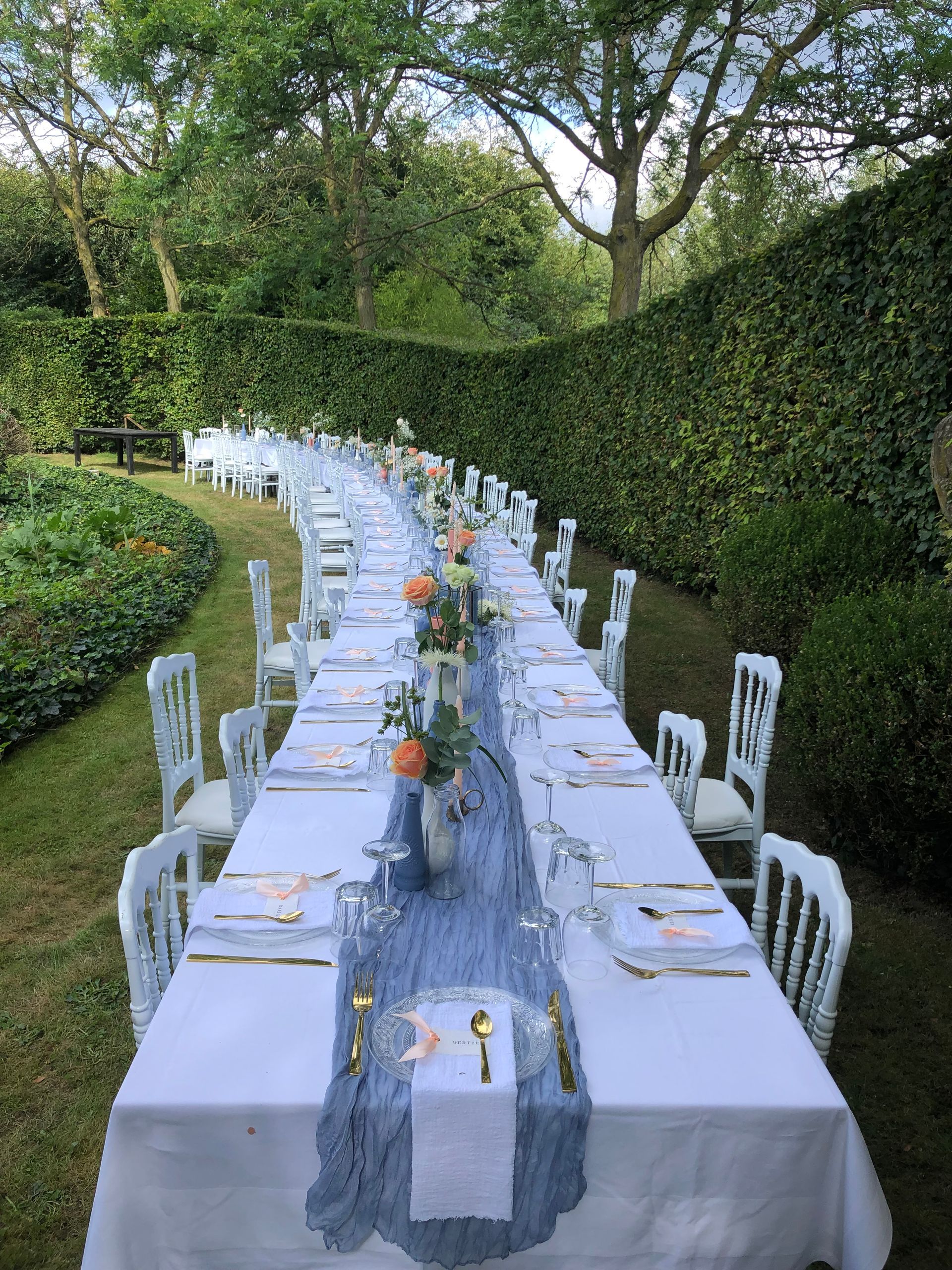 Lange eettafel voor buiten, gedekt voor een formele gelegenheid, met wit tafellinnen en een blauwe tafelloper.