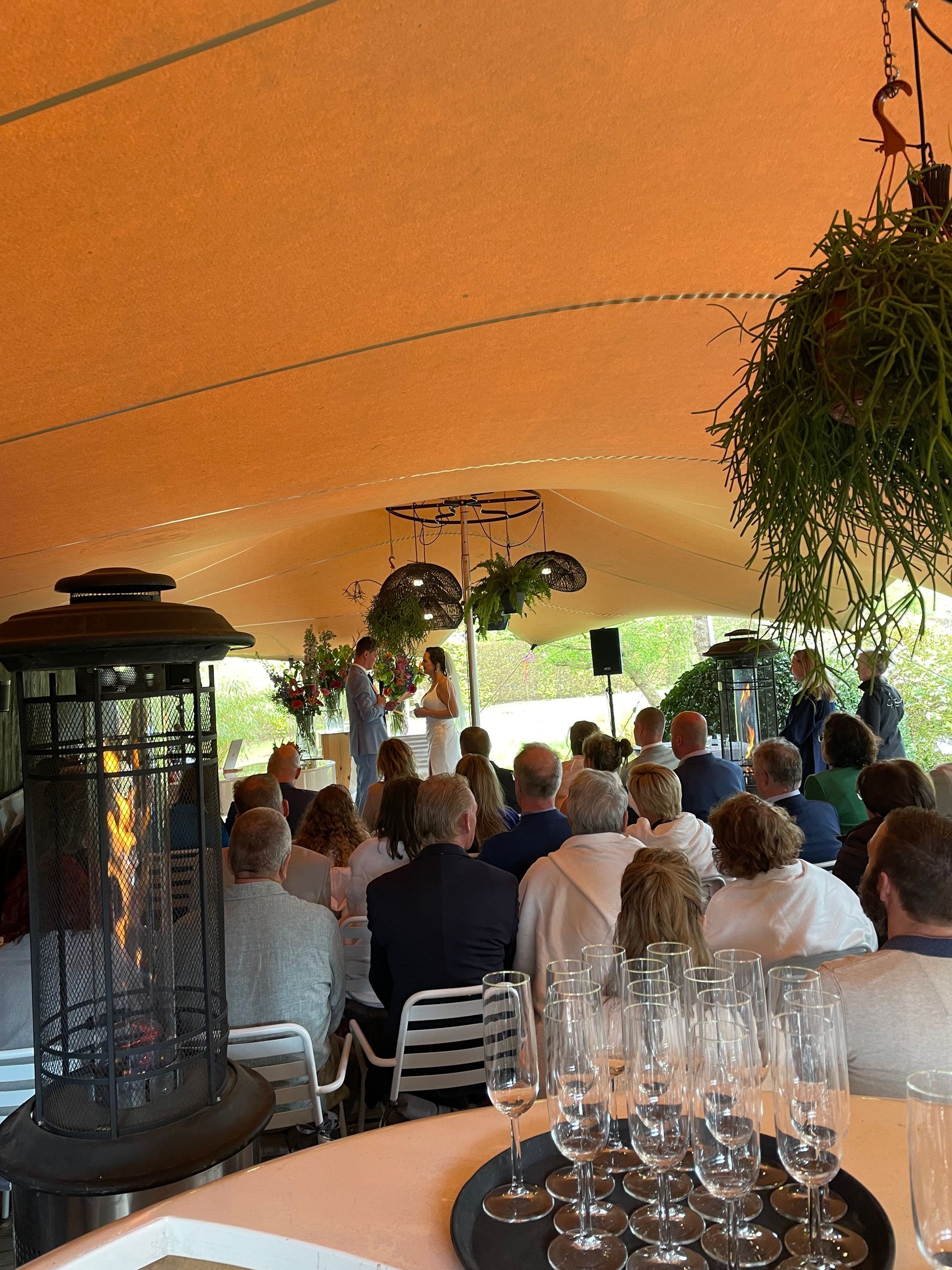 Huwelijksceremonie onder een tent. Gasten zitten, het bruidspaar staat bij het altaar, champagneglazen staan ​​op een tafel.