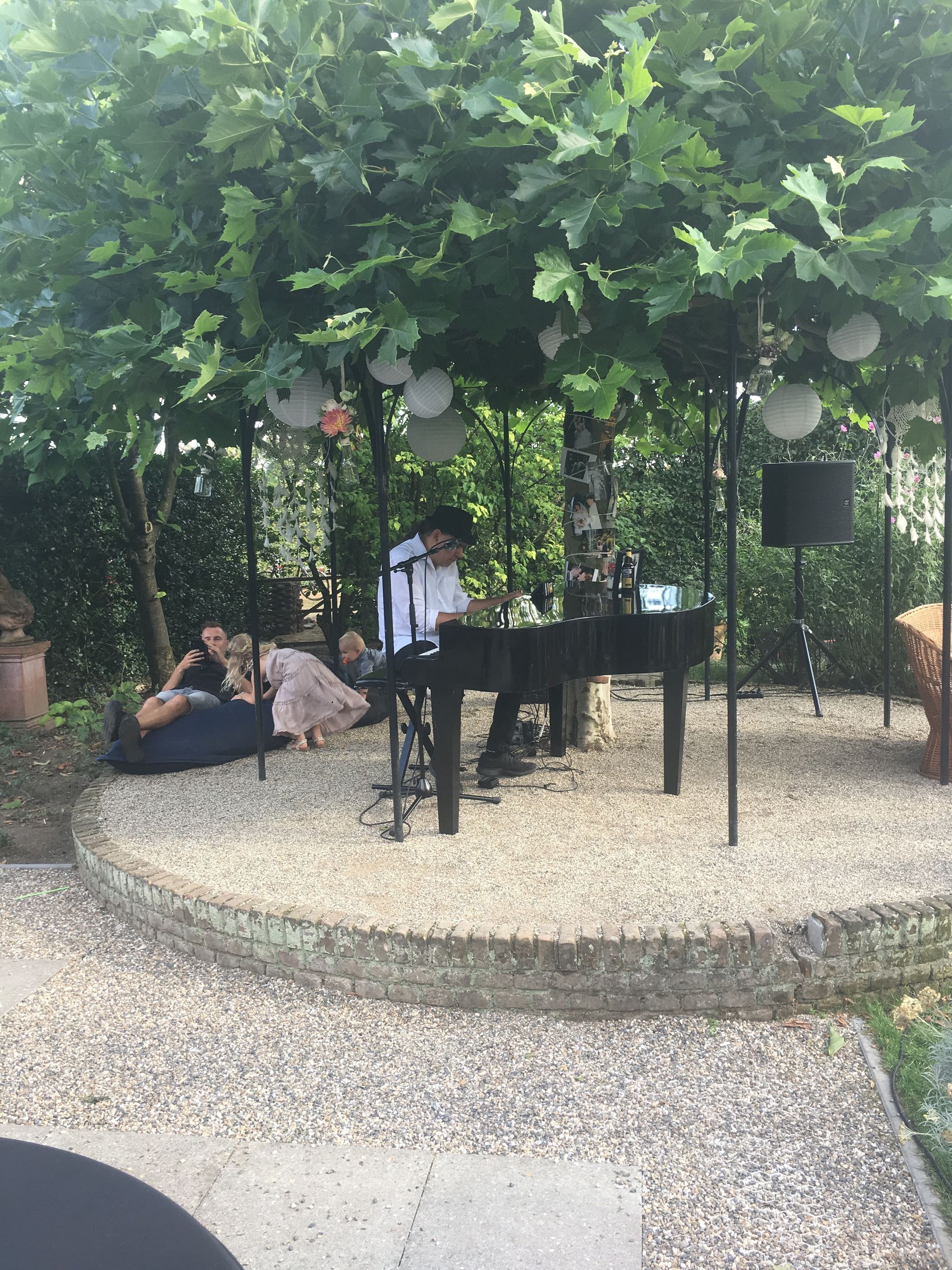 Een muzikant speelt piano in de buitenlucht onder een boom, met luisteraars op zitzakken.