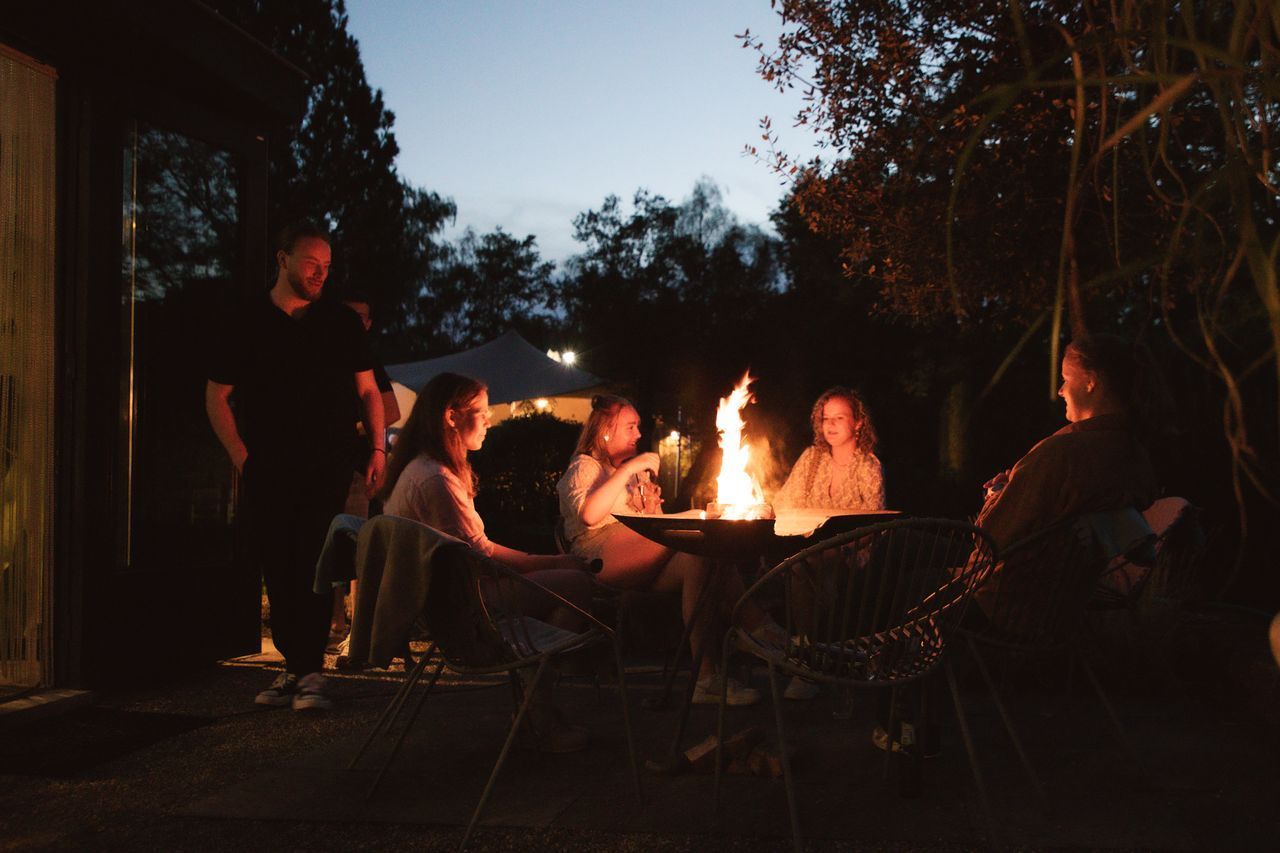 Mensen verzamelden zich rond een vuurplaats in de buitenlucht bij schemering. Bomen en een gebouw op de achtergrond.