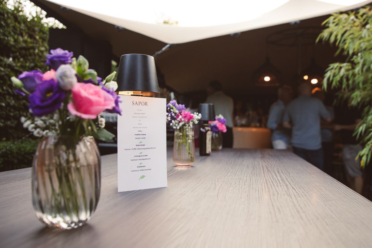 Bloemen, een menukaart en parfum op een houten tafel, met wazige mensen en een tent op de achtergrond.
