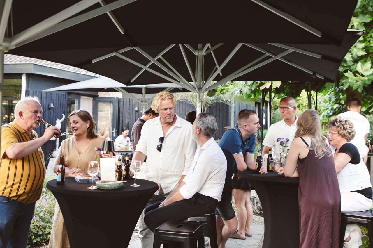 Een groep mensen die buiten onder een grote parasol gezellig samen zijn. Tafels met drankjes, een gebouw op de achtergrond.