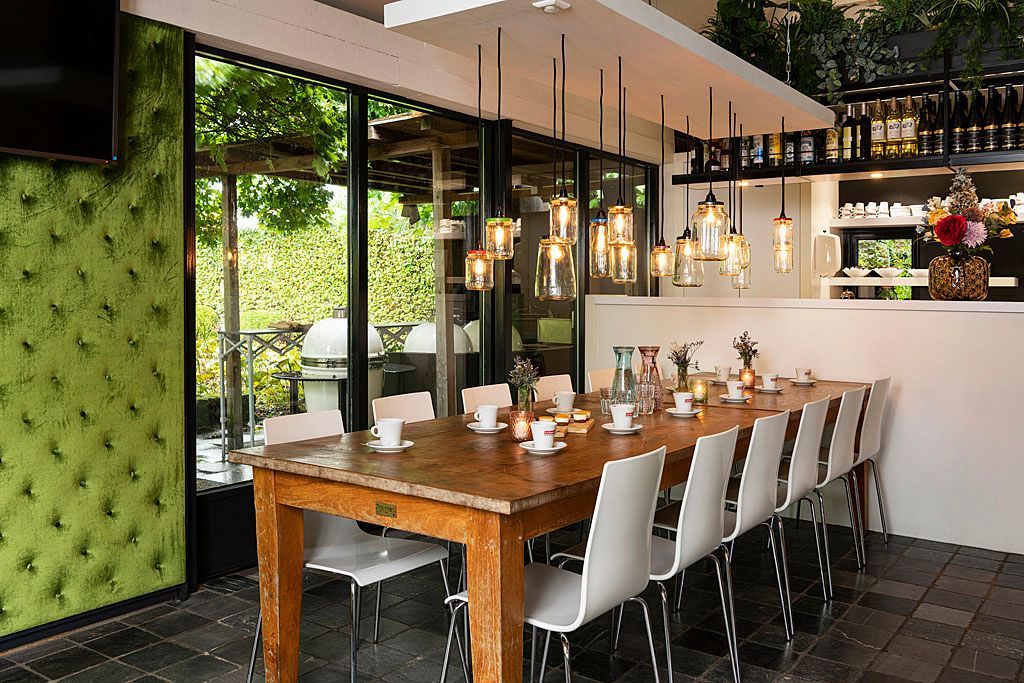 Een lange houten eettafel, gedekt voor gasten, met witte stoelen en hanglampen, staat naast een raam met uitzicht op groen.