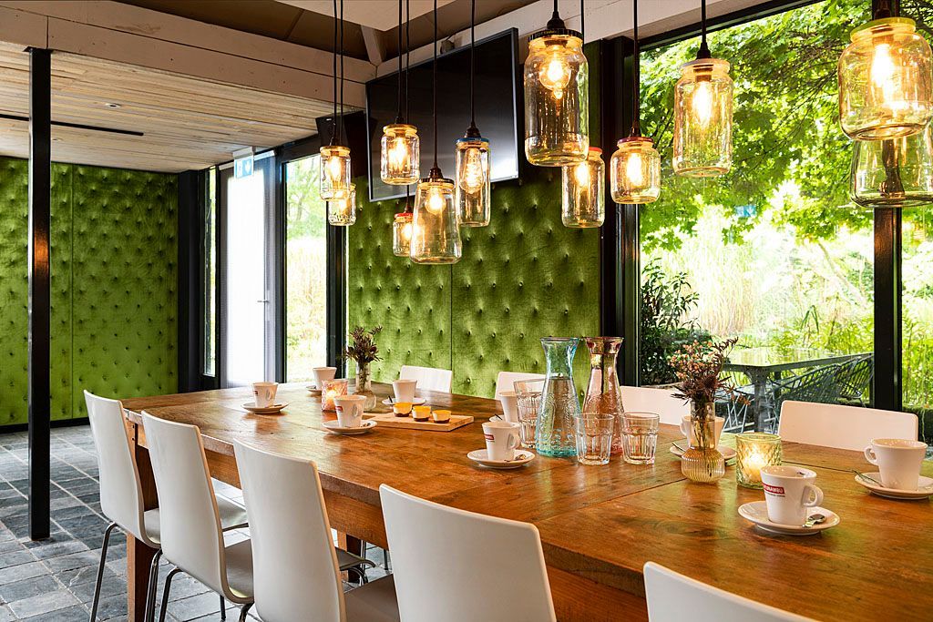 Eetkamer met lange houten tafel, witte stoelen, groene gecapitonneerde muur, hangende potlampen en uitzicht naar buiten.