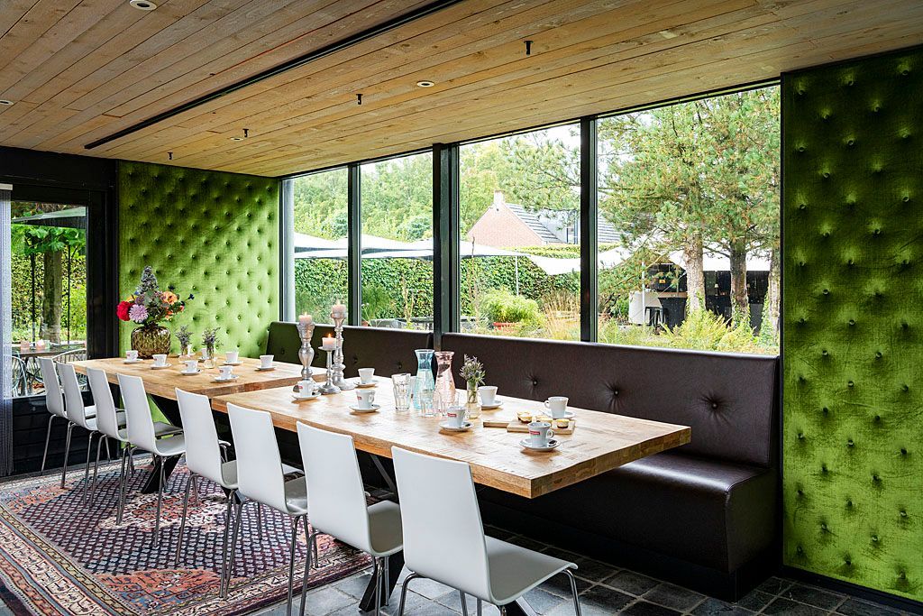 Een lange eettafel in een kamer met groene, gewatteerde muren en grote ramen die uitkijken op een tuin.