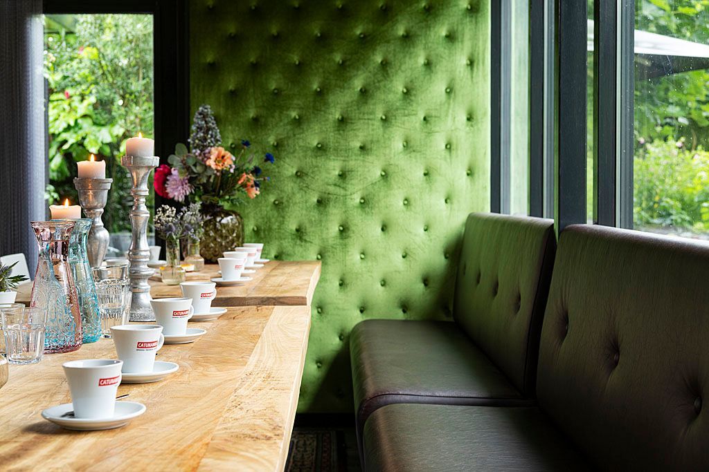 Een lange houten eettafel, gedekt met koffiekopjes, kaarsen en bloemen. Een groene, met gestoffeerde muur en een zitbank.