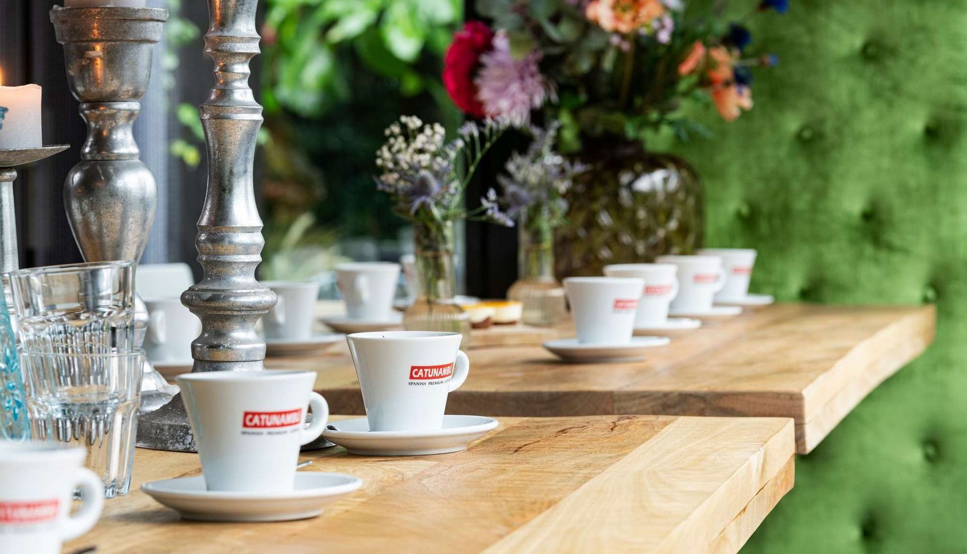 Espressokopjes op schoteltjes staan ​​op een houten tafel, naast een bloemstuk en zilveren kandelaars.