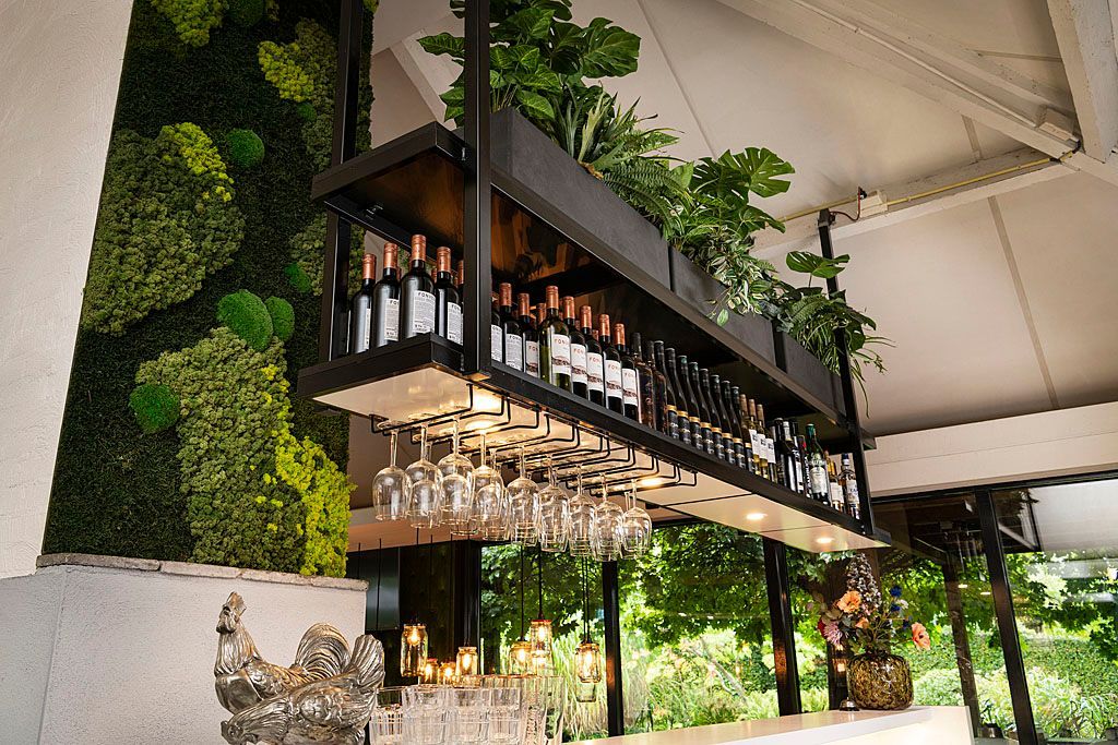 Bar met flessen en hangende glazen, groene wand, planten, buitenomgeving.