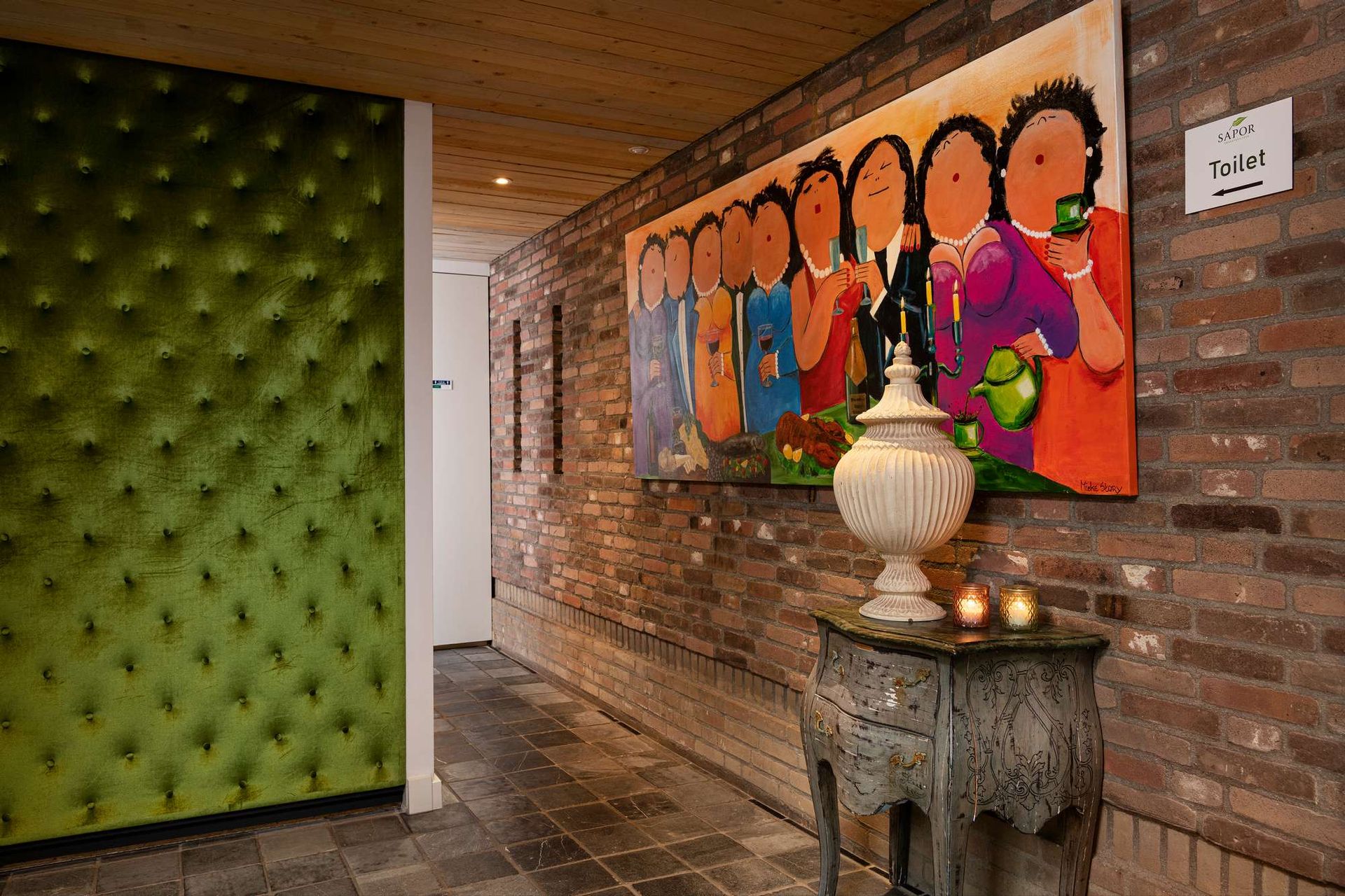 Hal met groene fluwelen wand, bakstenen muur, groot kleurrijk schilderij en decoratieve tafel met kaarsen.