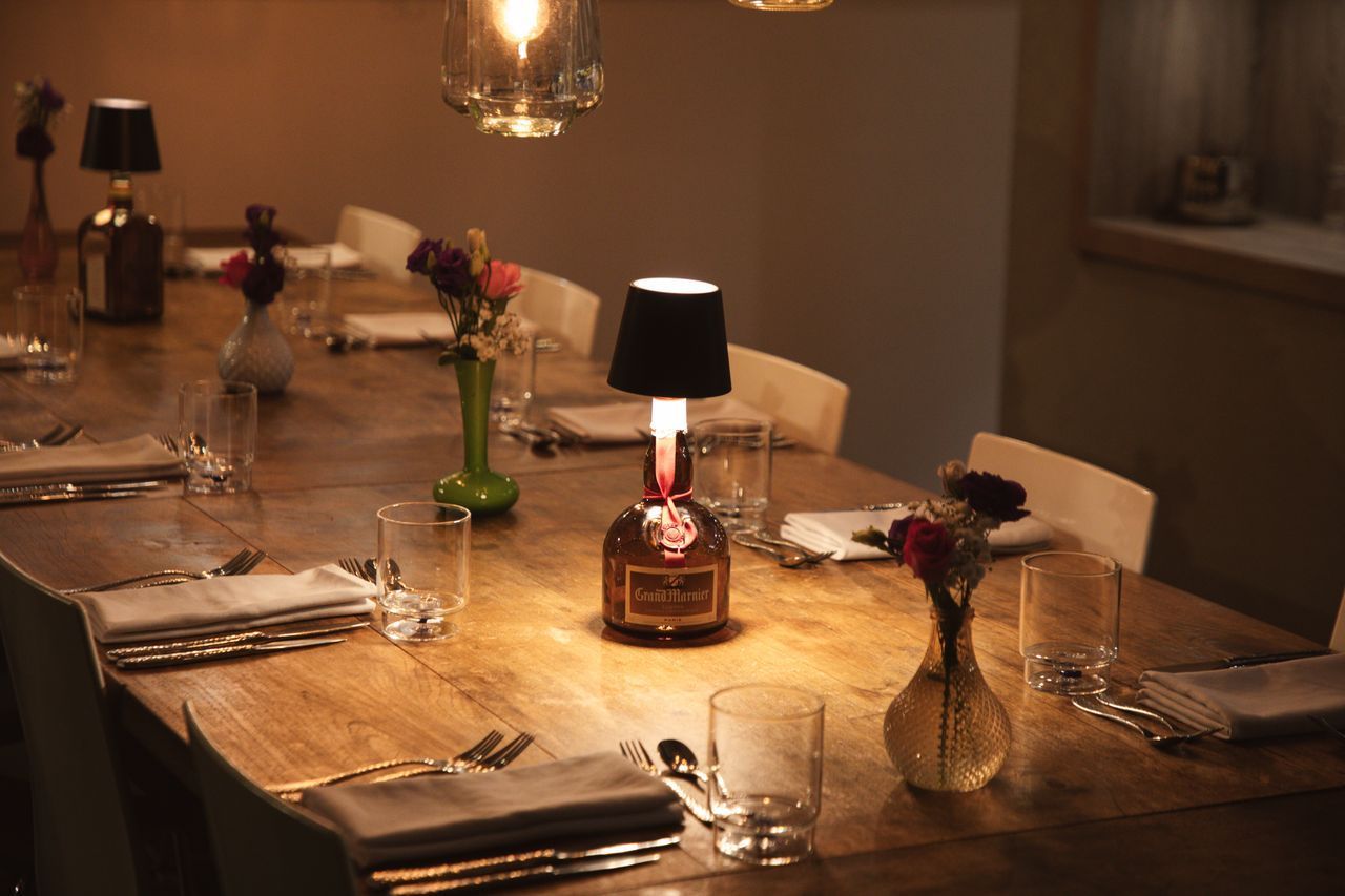 Een lange houten tafel gedekt voor het diner, met lampen gemaakt van flessen en bloemen, in een schemerig verlichte kamer.