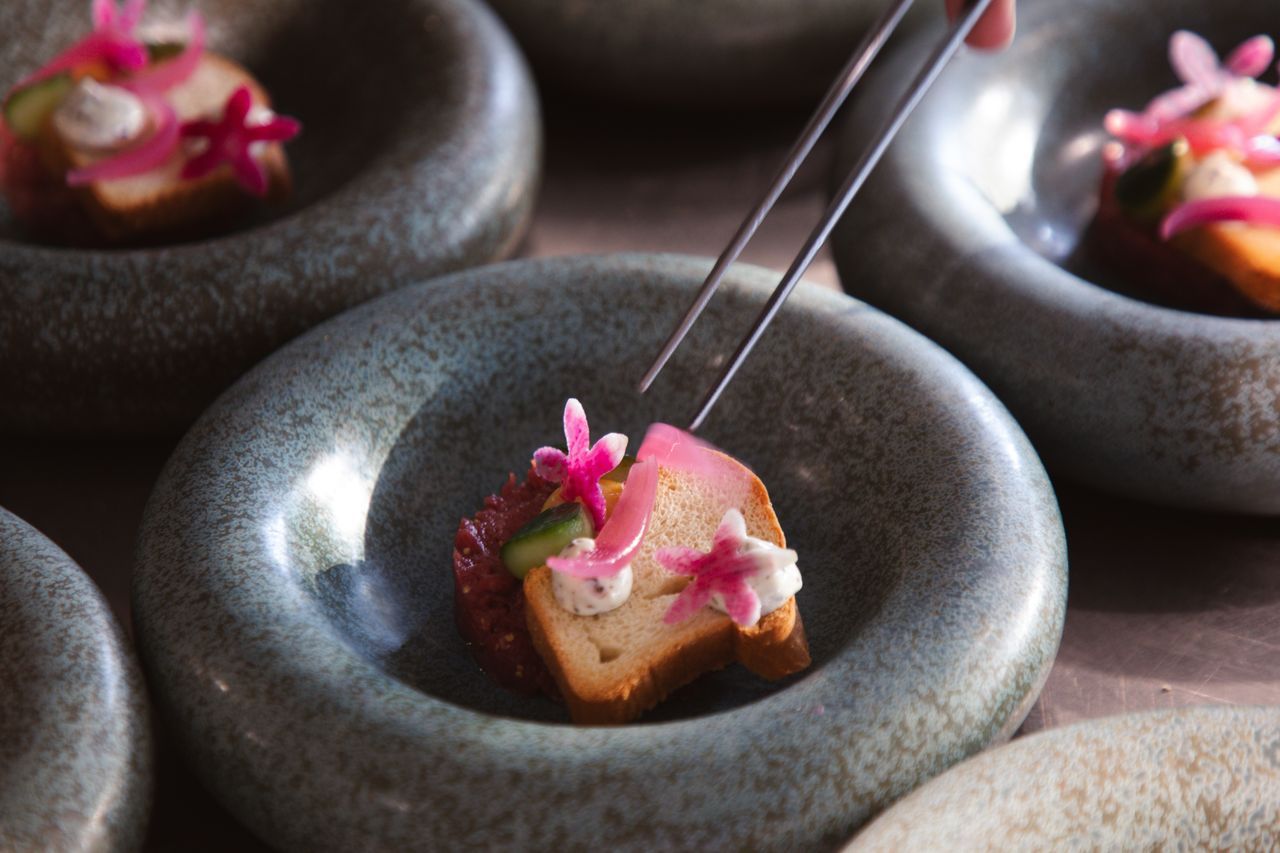De hapjes worden met een pincet gegarneerd en geserveerd in grijze stenen kommen met roze bloemen.