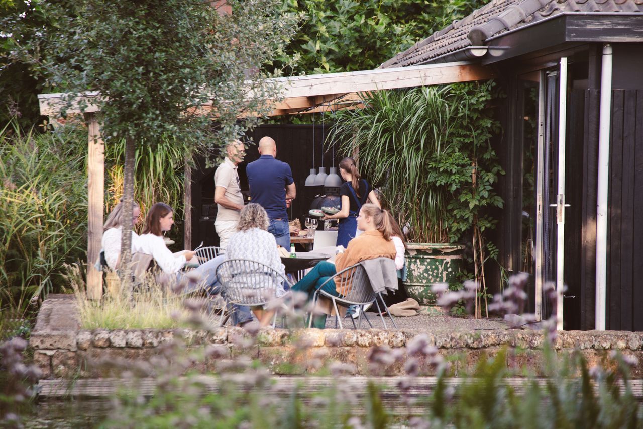 Een groep mensen had zich buiten verzameld, bij een vijver, onder een houten constructie en naast een klein gebouw.