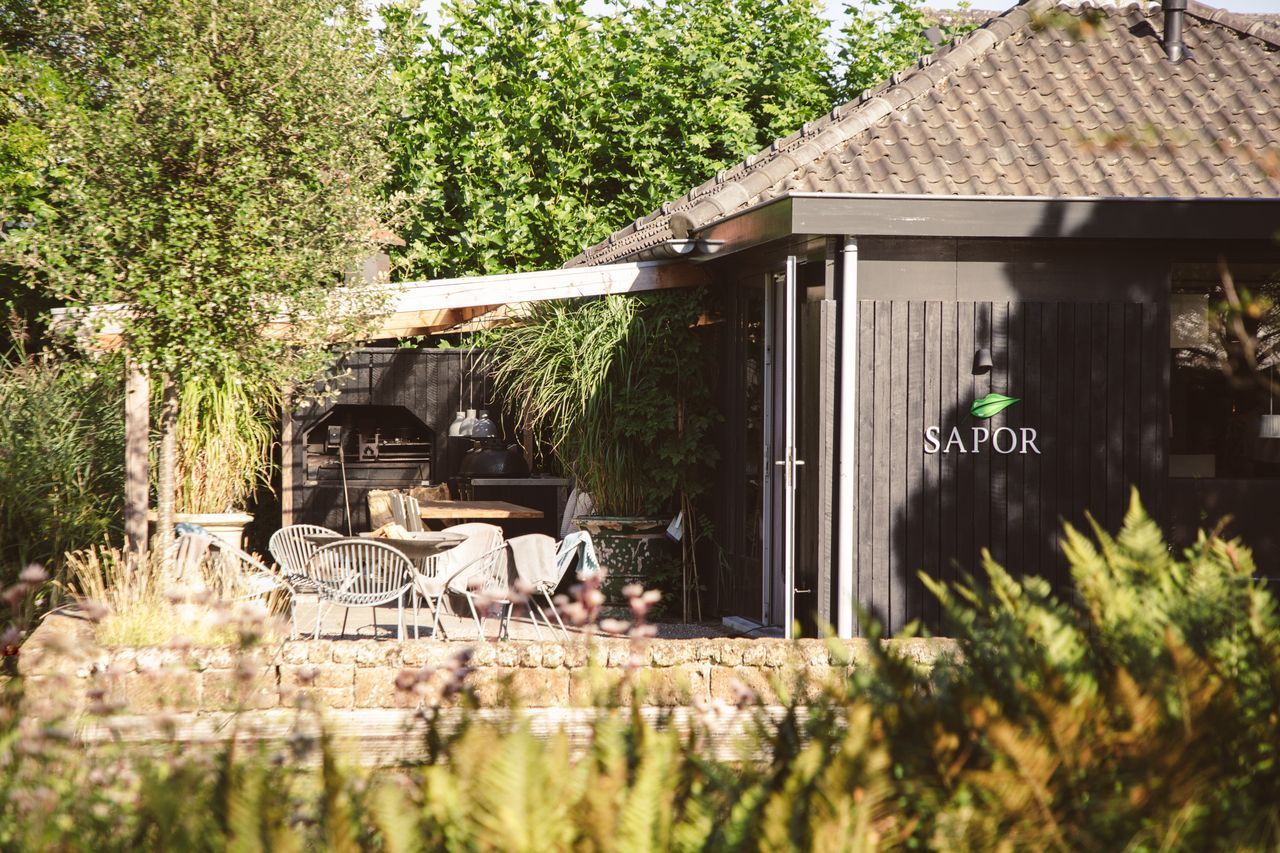 Buitengebouw met donkere gevelbekleding, pannendak, beplanting en een bord met de tekst 