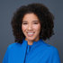 A woman with curly hair is wearing a blue jacket and smiling.