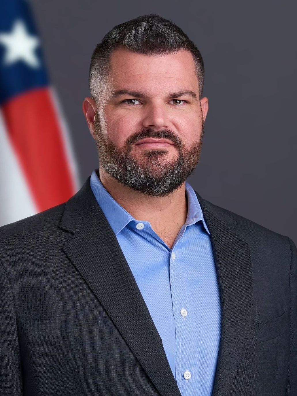 A man in a suit and blue shirt is standing in front of an american flag.