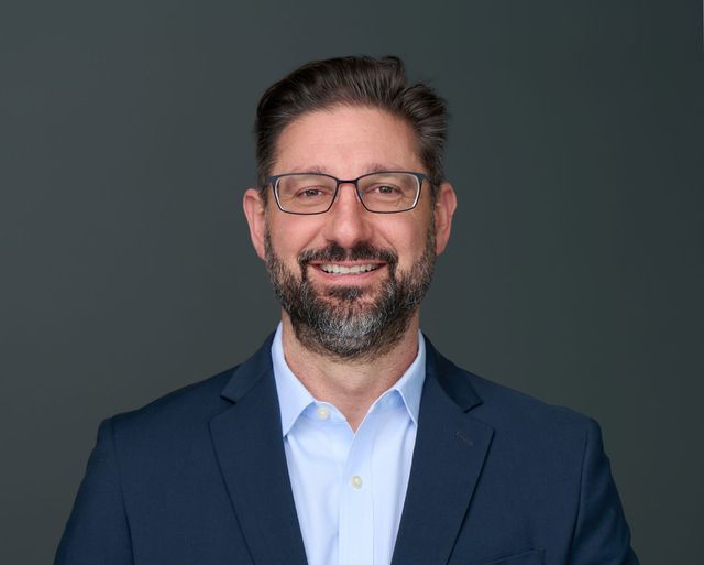 Smiling man in glasses and navy blazer against a dark gray background