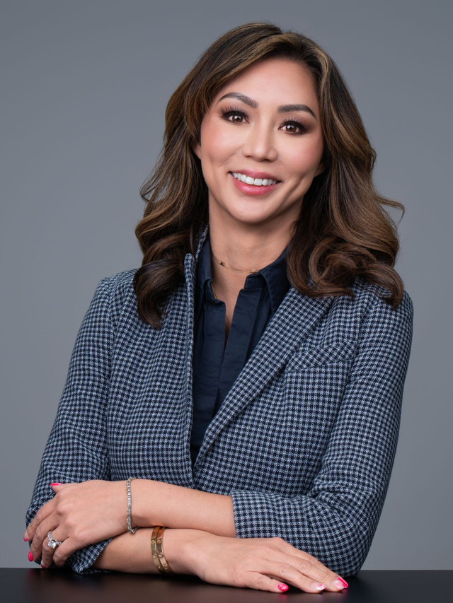 Woman in a blue blazer smiling with hands folded against a gray background