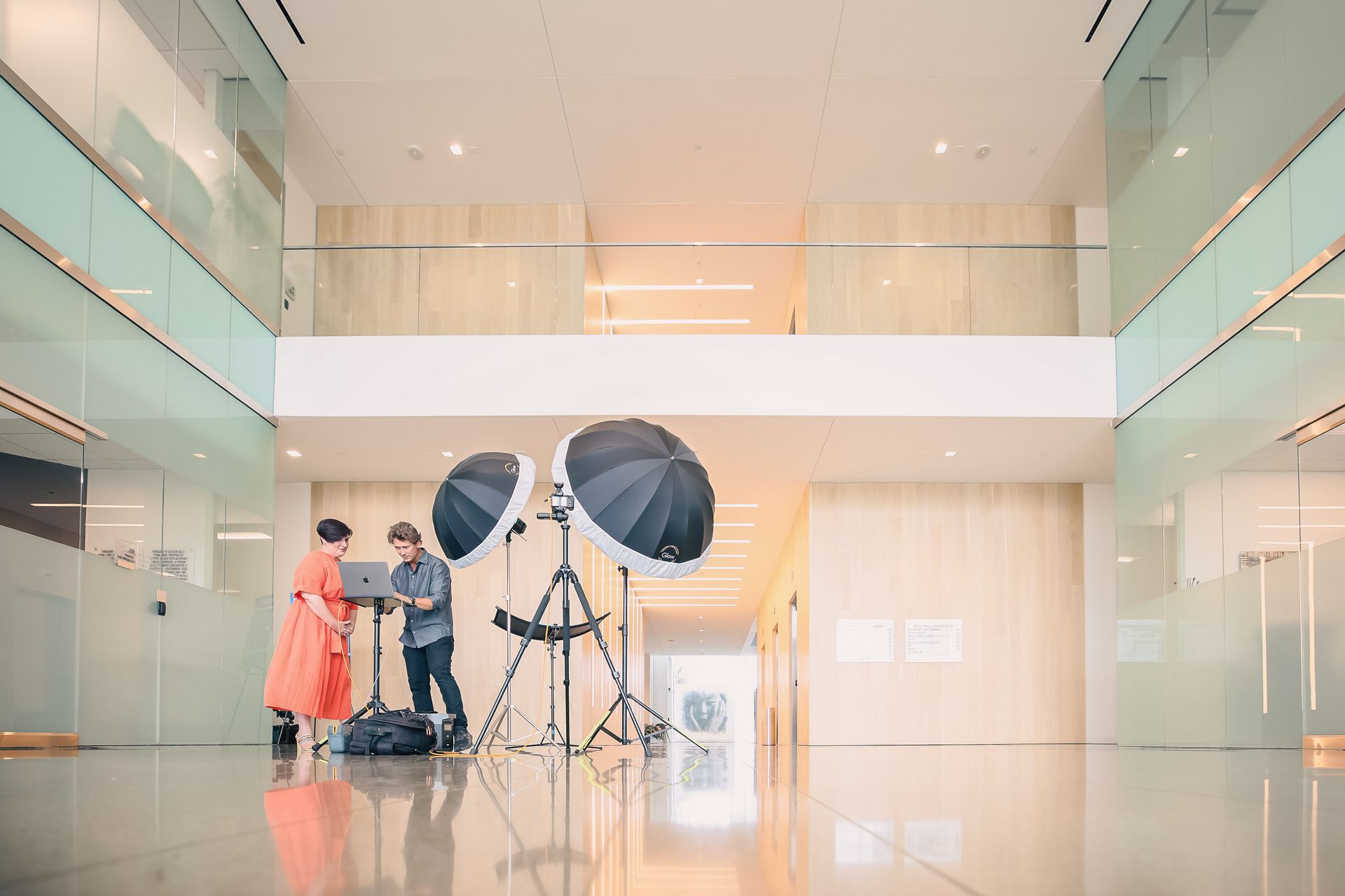 A man and a woman are standing in a hallway with umbrellas and a laptop.