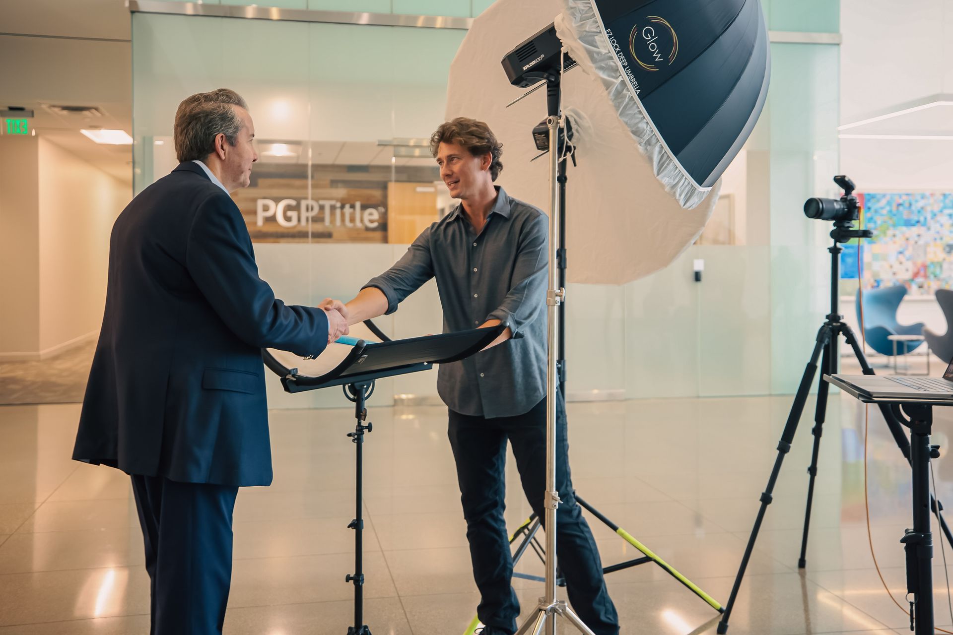 Two men are shaking hands in front of a camera in a room.