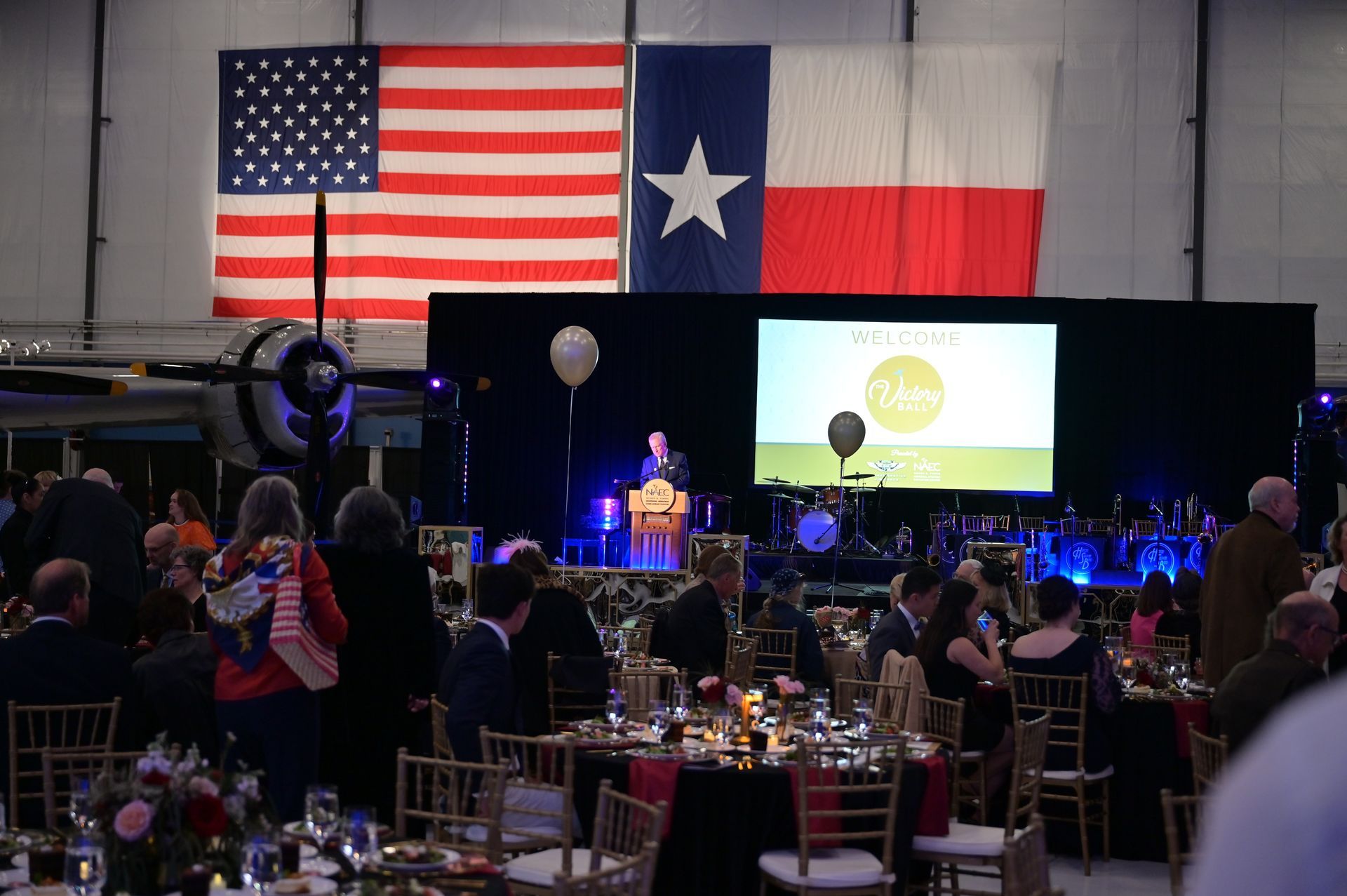 A large group of people are sitting at tables in front of a large american flag and a large texas flag.