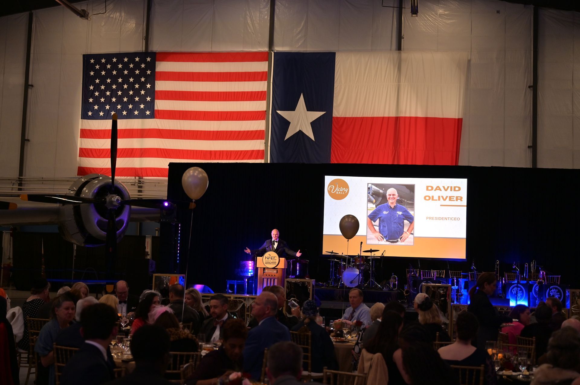 A man is giving a speech at a dinner in front of an american flag and a texas flag.