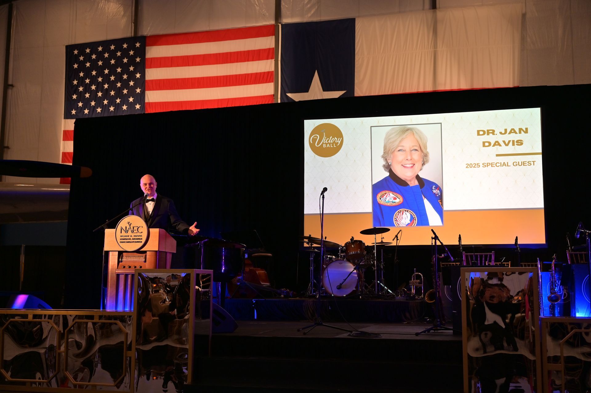 A man stands at a podium in front of a large screen with a picture of a woman on it