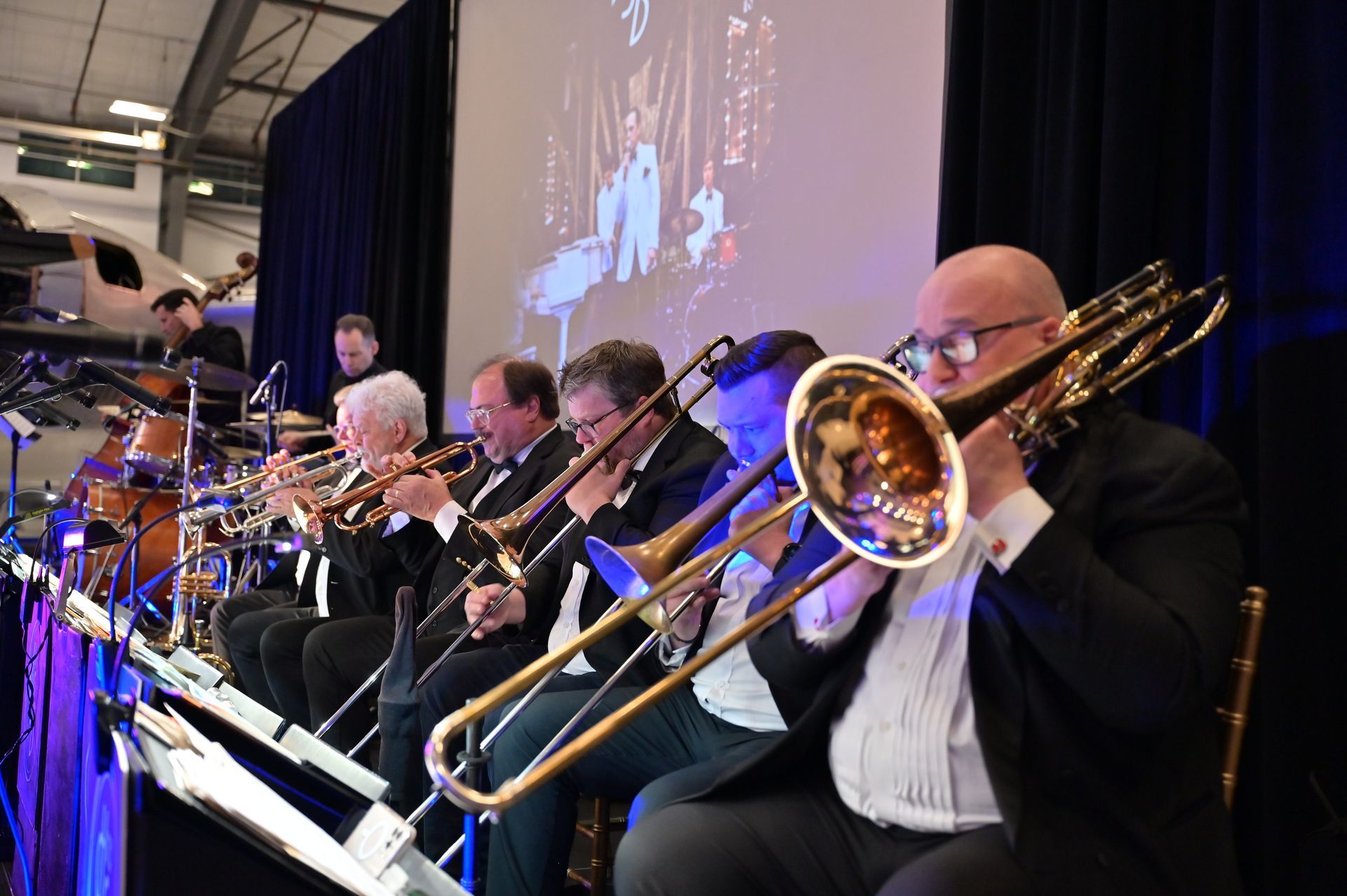 A group of men are playing trombones in front of a large screen.
