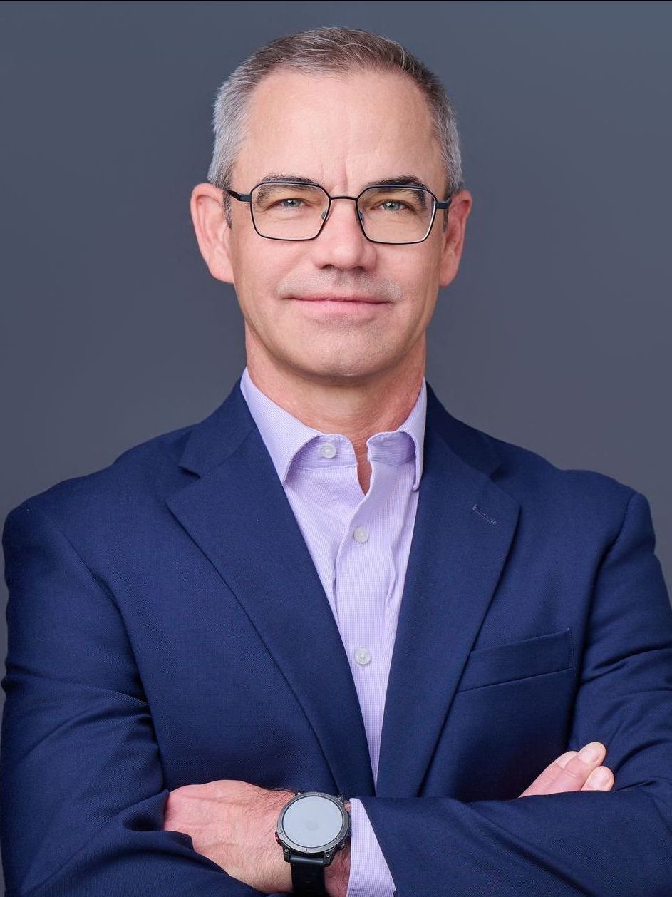 A man in a blue suit and glasses is standing with his arms crossed.