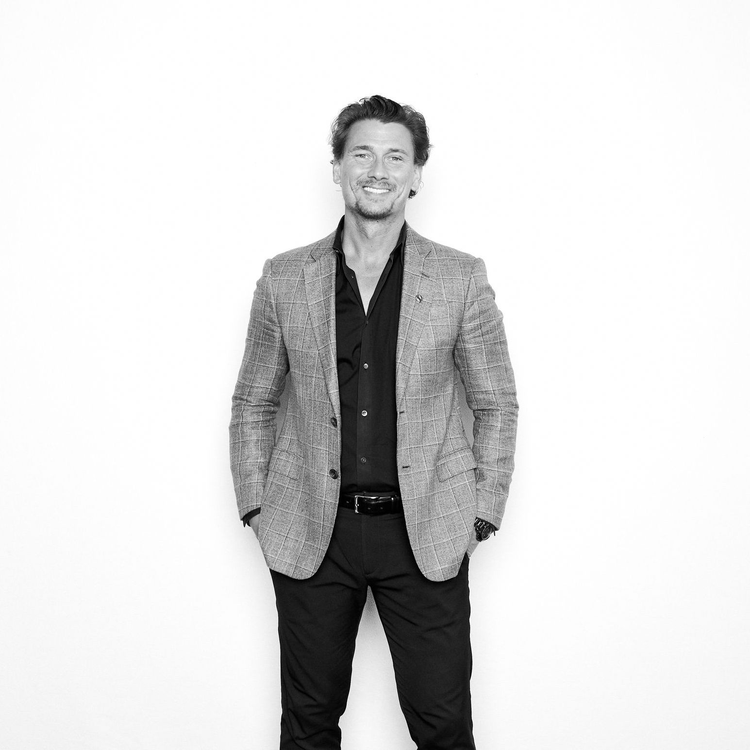 A man in a suit is standing in front of a white wall with his hands in his pockets.