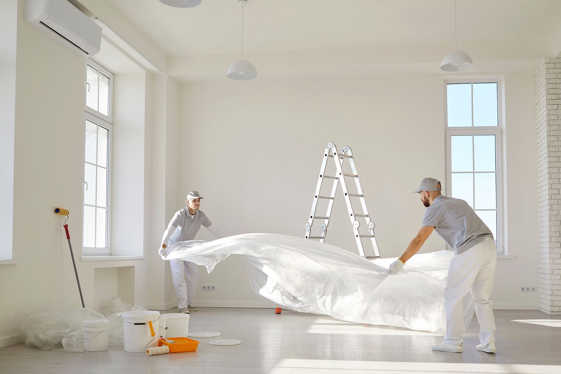Two painters in white coveralls spreading a large white sheet in a bright, empty room with a ladder.