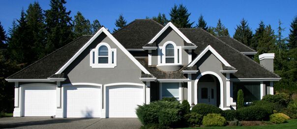 Large white suburban house with dark roof, three-car garage, and manicured front lawn under a blue sky