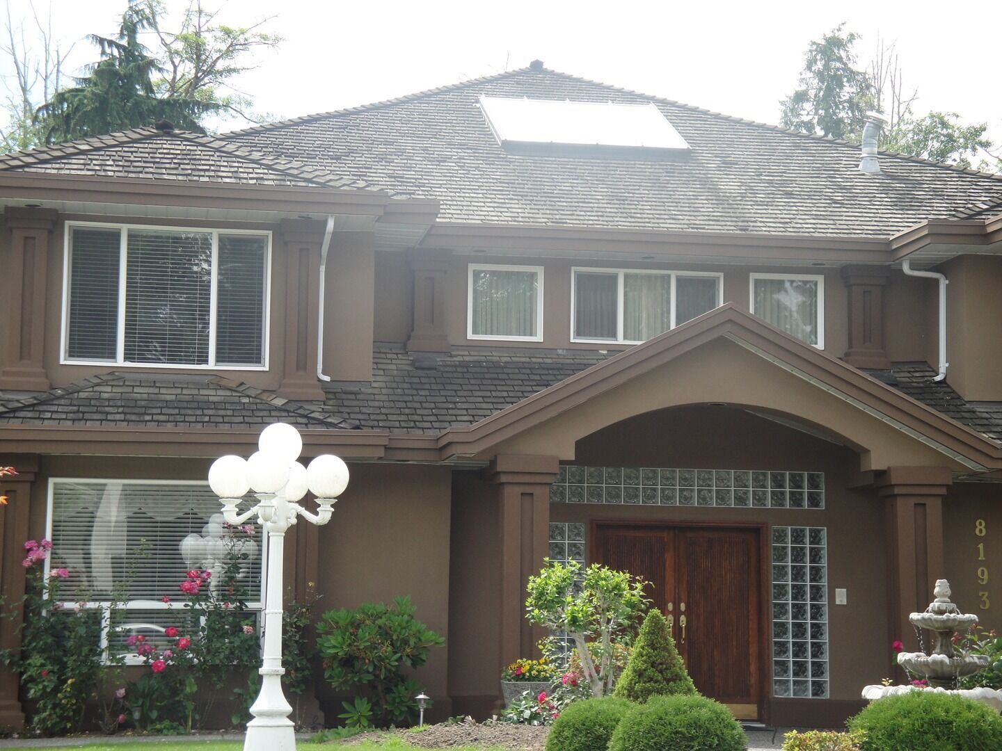 Two-story brown house with arched entry, white lamp post, and flowered front yard