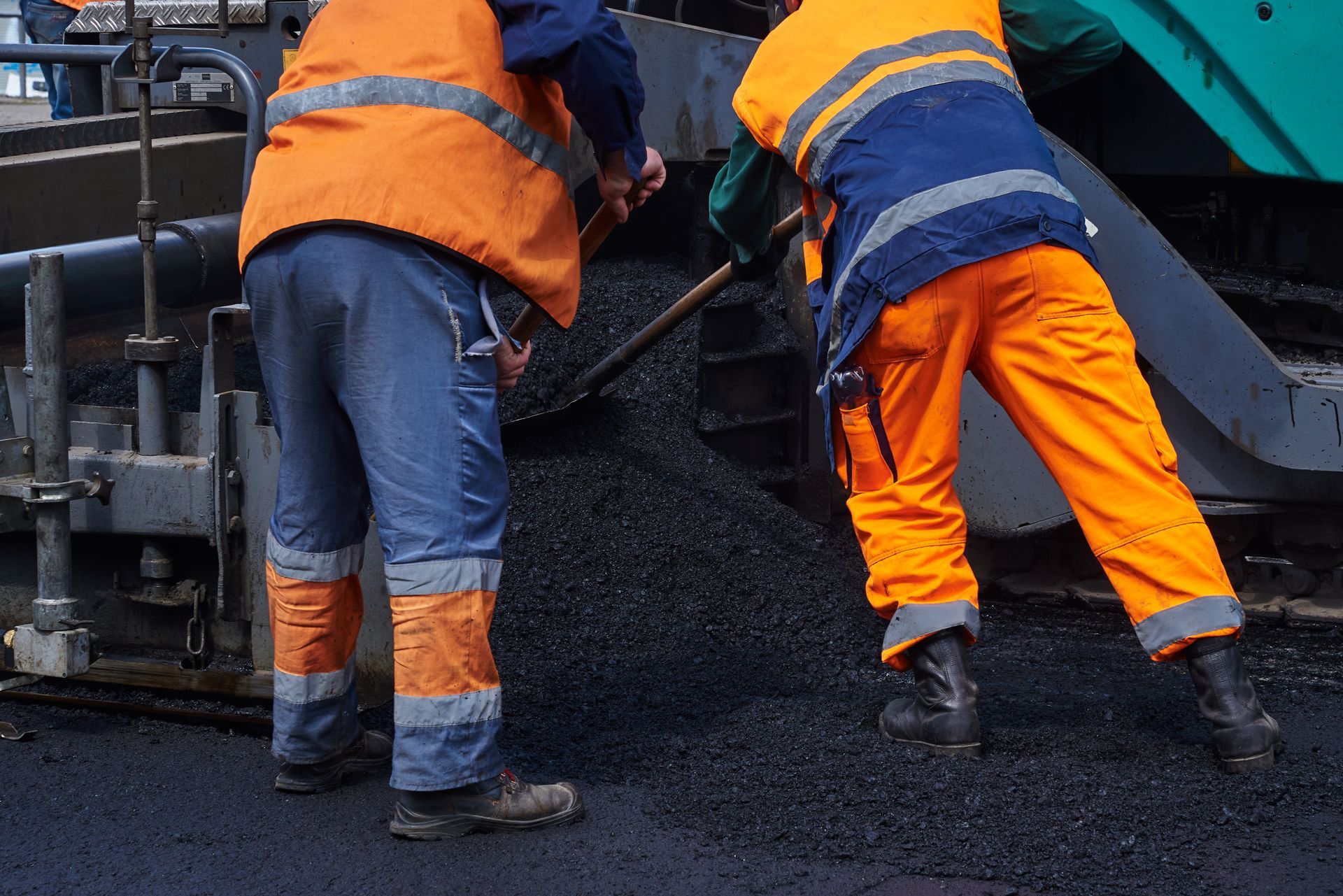 Two construction workers in high-visibility orange vests and blue pants spread asphalt on a road.