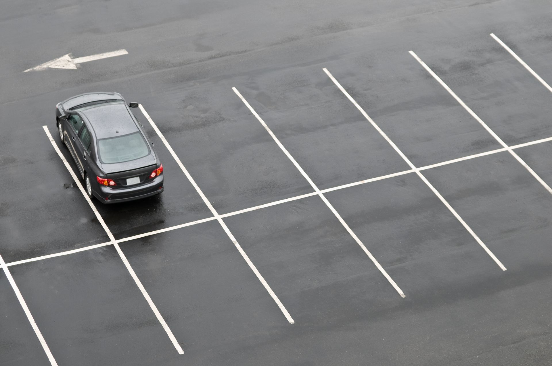 A dark car parked in the first spot of a nearly empty, wet asphalt parking lot with white painted lines and an arrow.
