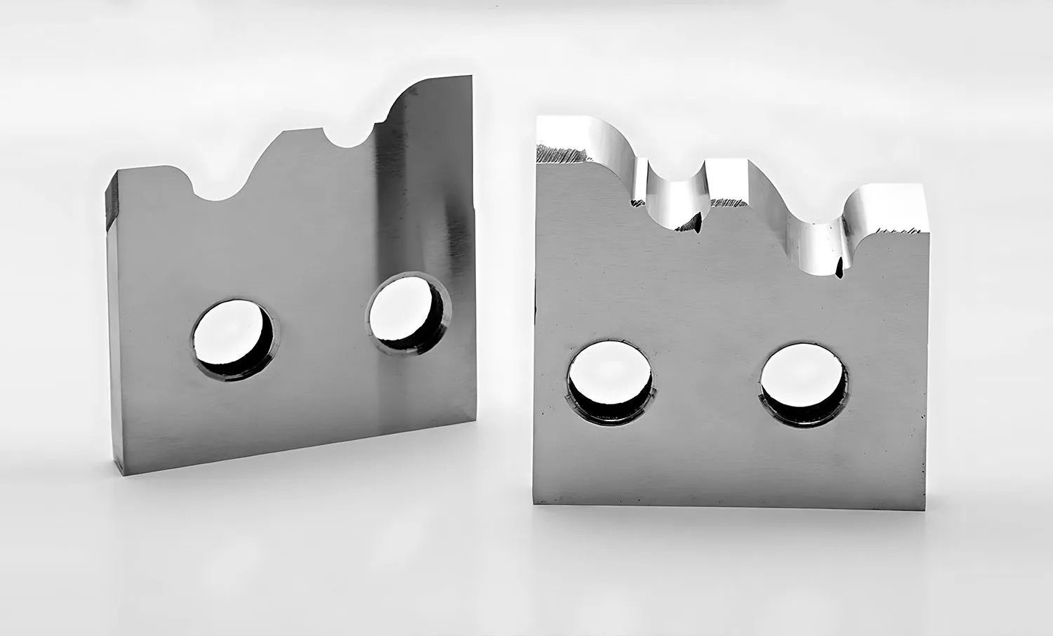 Two shiny, metal, notched rectangular parts with holes and a wavy top edge.