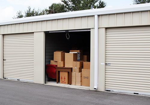 Storage unit with beige roll-up door open, revealing cardboard boxes and small furniture. Two closed units on either side.