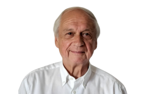 Un homme âgé aux cheveux blancs sourit, portant une chemise blanche boutonnée sur un fond blanc.