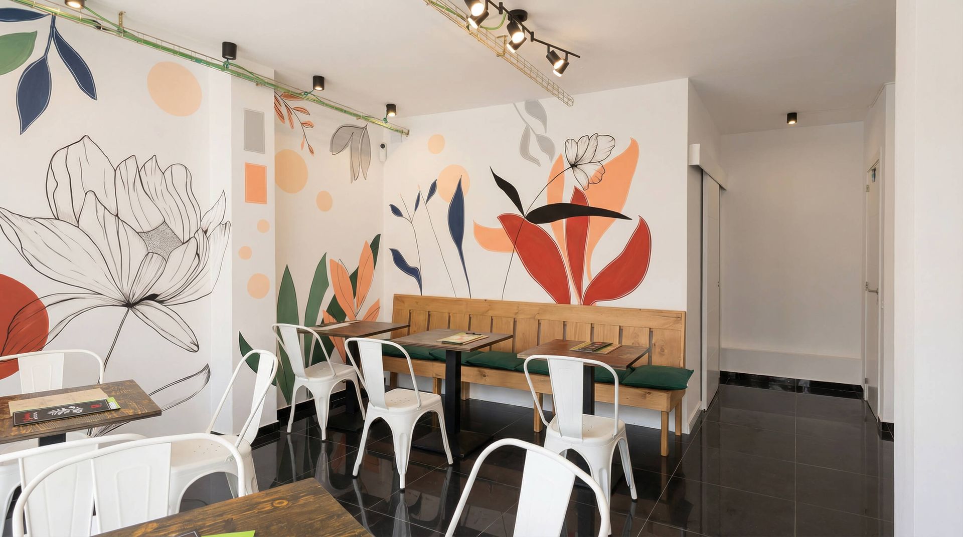 Interior de cafetería con sillas blancas, mesas de madera y mural floral.