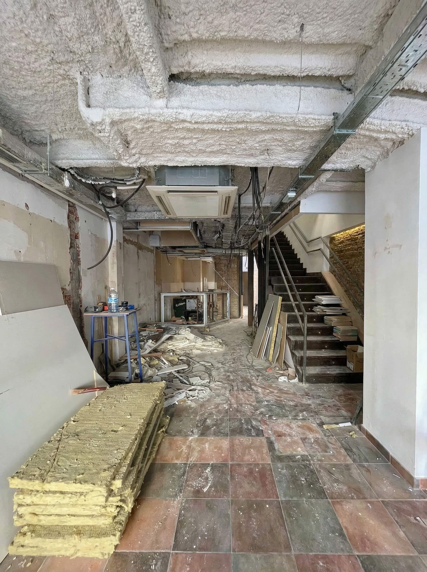 Vista interior de una obra en construcción. Escombros cubren el suelo, techo expuesto con tuberías y conductos