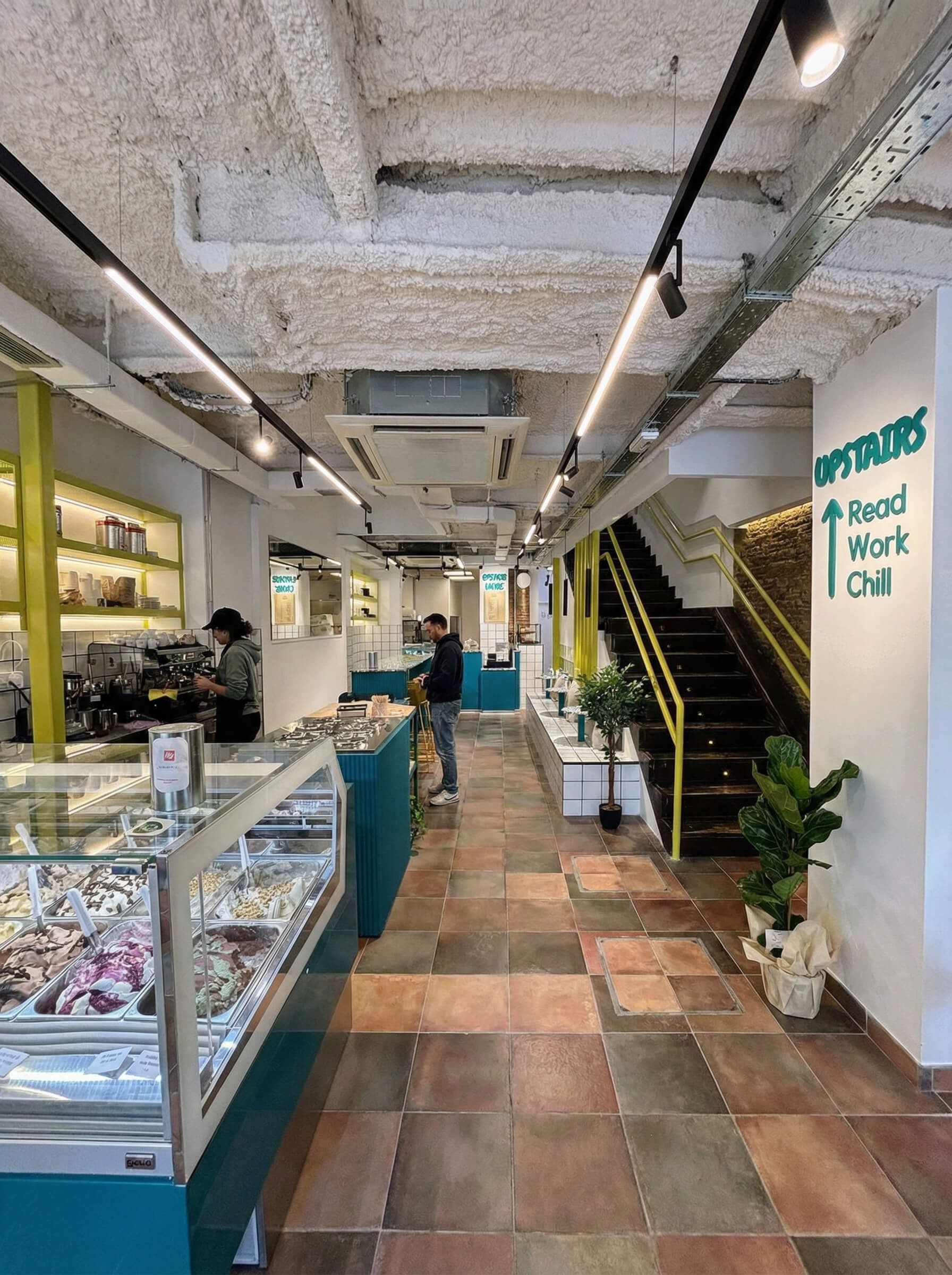 Interior de una cafetería con vitrinas de cristal, clientes y escaleras que conducen hacia arriba.