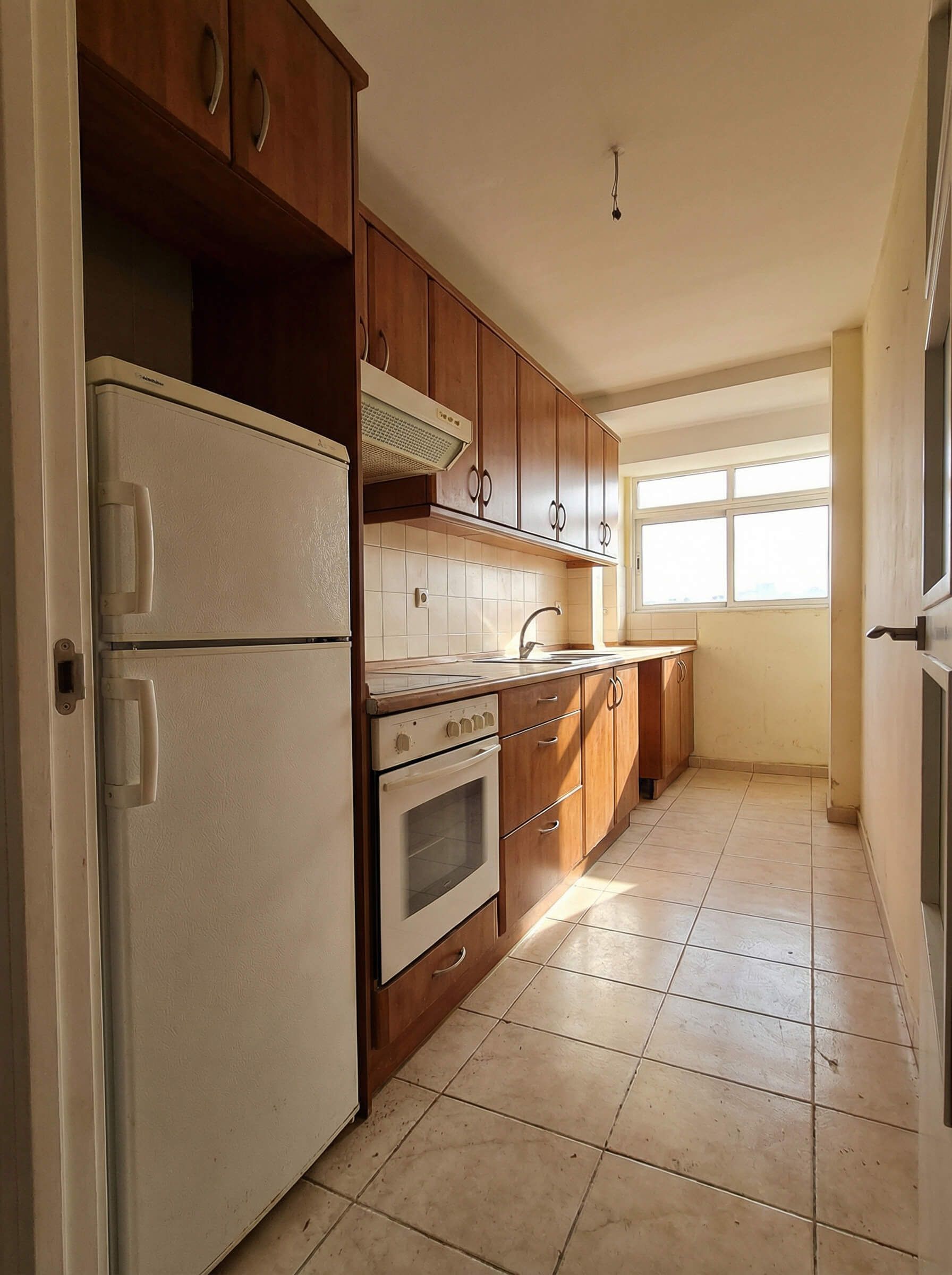 Cocina estrecha con armarios de madera, nevera y horno. La luz del sol entra a raudales por una ventana.