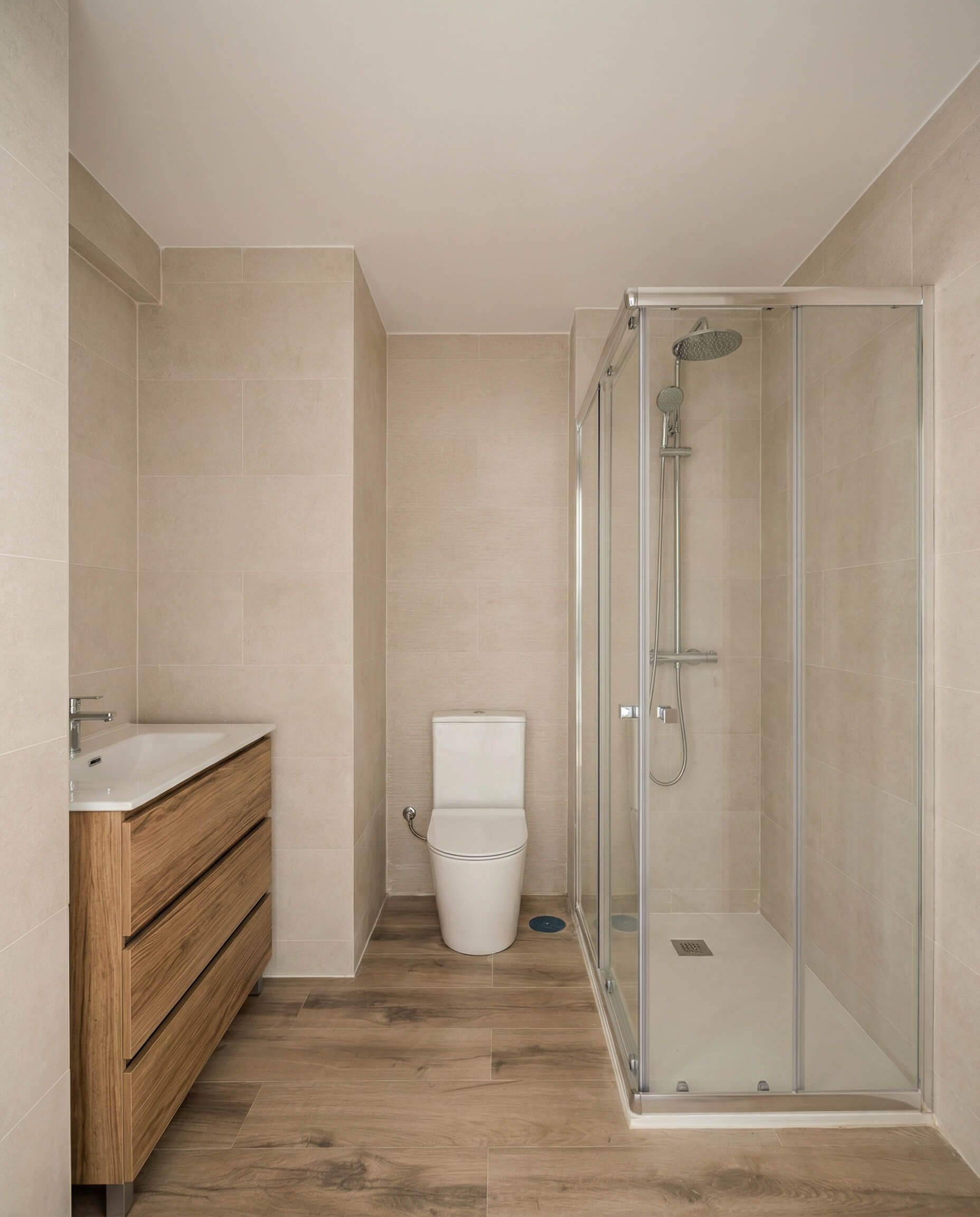 Baño con tocador de madera, inodoro blanco, ducha de cristal y paredes y suelos de azulejos beige.