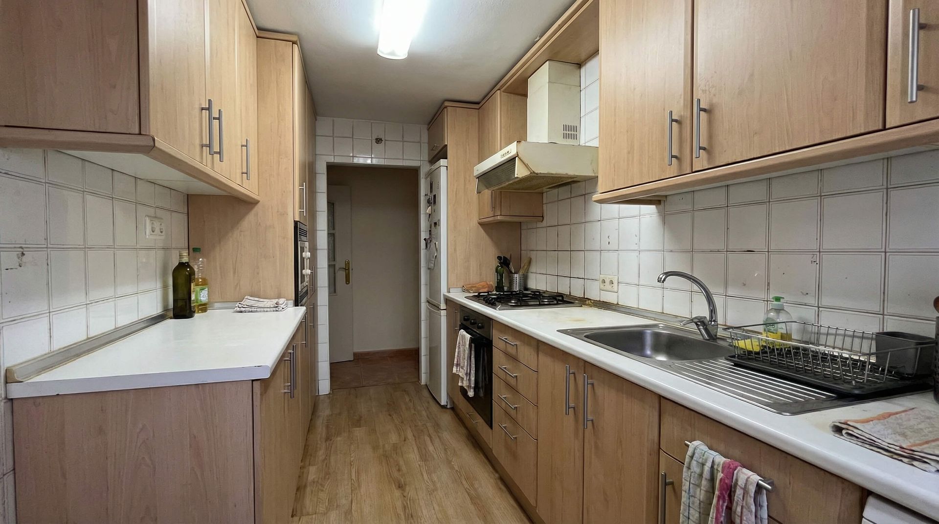Cocina estrecha con gabinetes de madera clara, encimeras blancas y protector contra salpicaduras de azulejos.