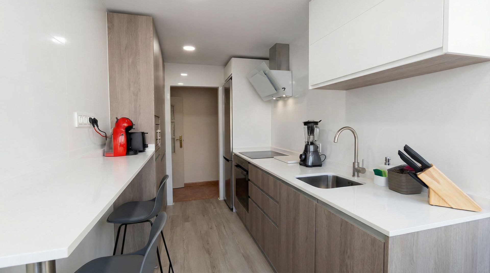 Una cocina moderna y estrecha con encimeras blancas y gabinetes de madera, asientos de barra