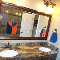 A double-vanity bathroom featuring a large framed mirror, dark wood cabinets, marble countertops, and orange/blue towels.