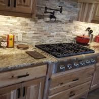 A modern kitchen stove with a black pot filler faucet on a light stone backsplash, light wood cabinets, and granite top.