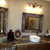 A bathroom vanity with a zebra-print framed mirror, granite countertop, vessel sink, and three-light wall fixture.