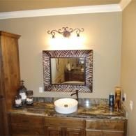 A bathroom vanity with a zebra-patterned mirror, dark stone countertop, vessel sink, and warm-toned wood cabinetry.