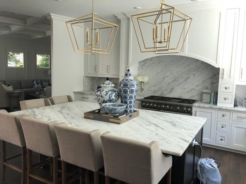 A bright kitchen with a large marble island, four beige bar stools, gold geometric pendant lights, and blue-white vases.