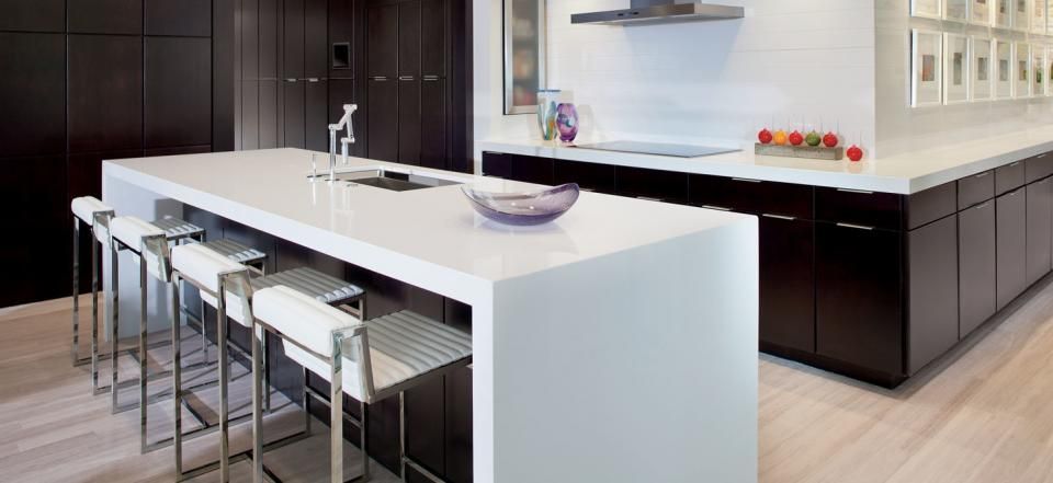 Modern kitchen with a white waterfall island, bar stools, and dark wood cabinetry on light hardwood floors.